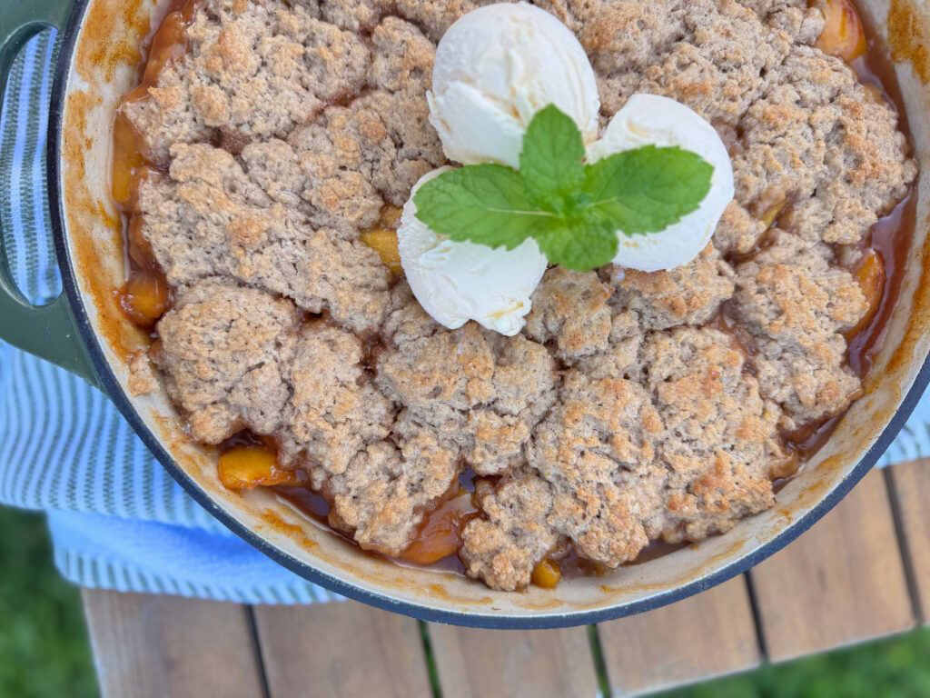 Baked homemade buttermilk peach cobbler in a green-handled baking dish, topped with golden-brown biscuit-style topping, three scoops of vanilla ice cream, and a sprig of fresh mint