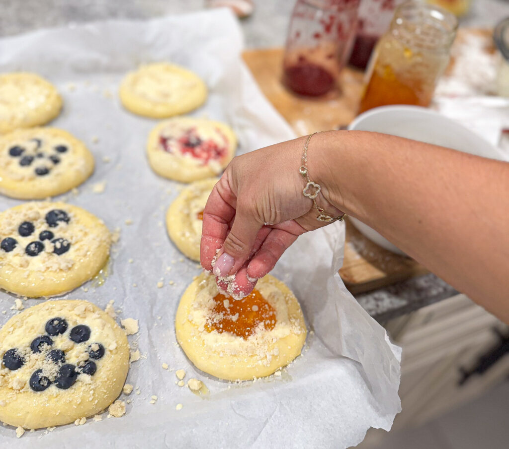 A hand sprinkles crumb topping over homemade sourdough kolaches filled with apricot jam on a parchment-lined baking sheet. Blueberry and strawberry-filled kolaches are also visible in the background, along with jars of fruit jam.