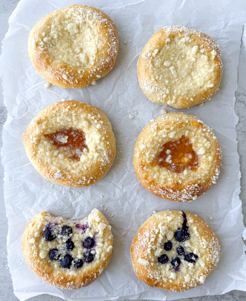 Six baked sourdough kolaches on parchment paper, topped with crumb streusel and filled with cream cheese, apricot jam, and blueberry. One blueberry kolache has a bite taken out, showing the soft fluffy texture inside.