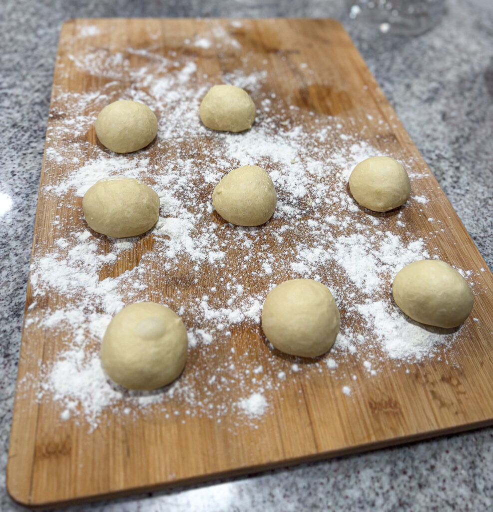 Small sourdough kolache dough balls resting on a wooden board dusted with flour, ready to rise before filling.