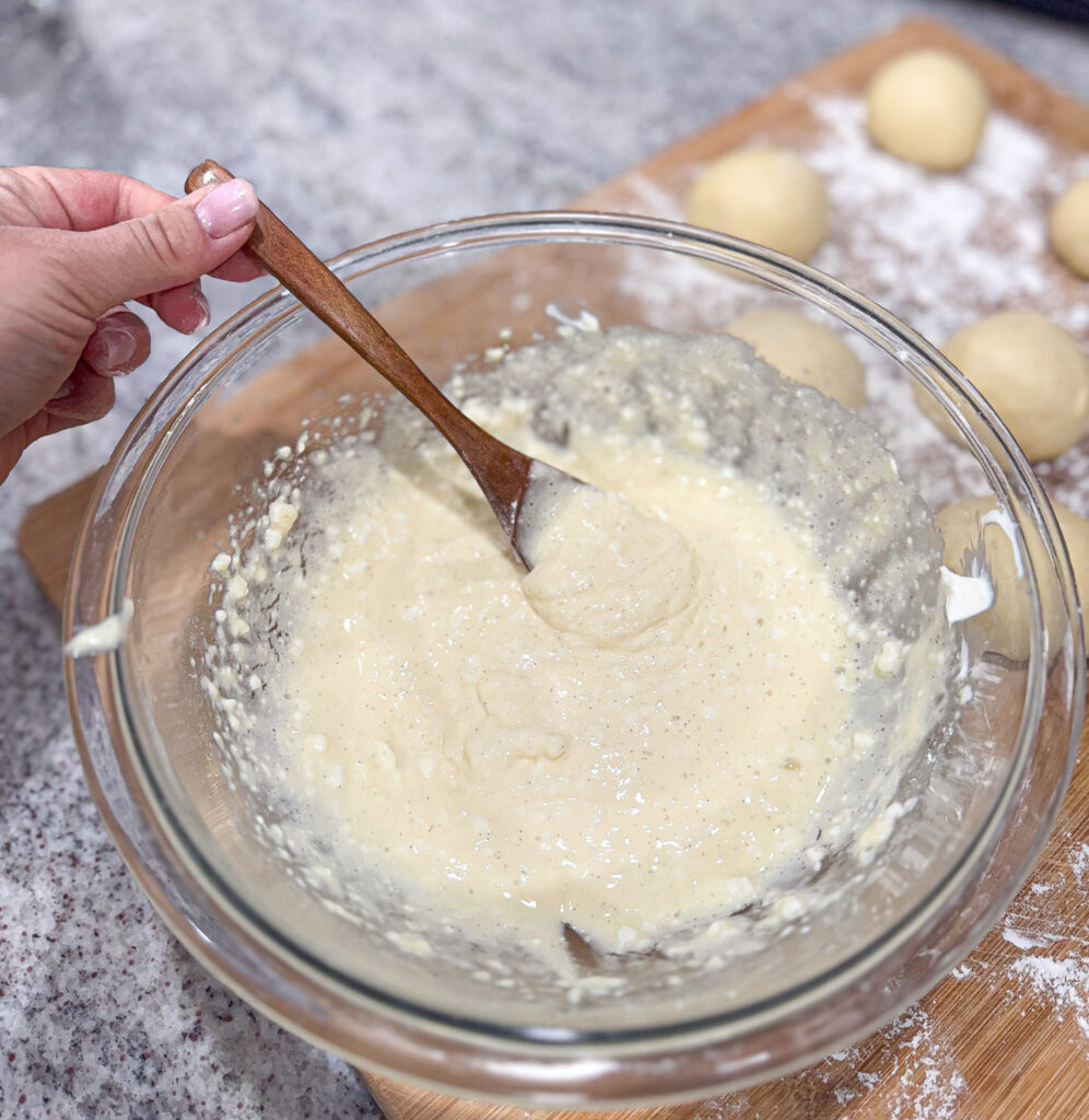 A glass bowl filled with sweet cream cheese filling being stirred with a wooden spoon, with sourdough kolache dough balls resting on a floured board in the background.