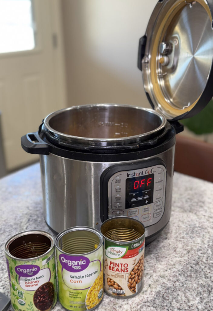 An Instant Pot sits open on a kitchen counter with the display showing "OFF." In front are three opened cans: dark red kidney beans, whole kernel corn, and pinto beans, ready to be used in a recipe.