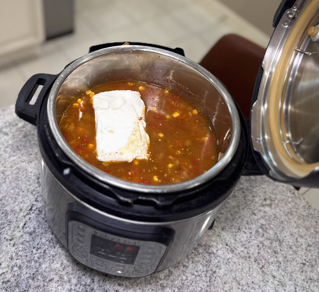 A close-up of an Instant Pot filled with a broth mixture of beans, corn, and tomatoes, topped with a block of cream cheese before cooking.