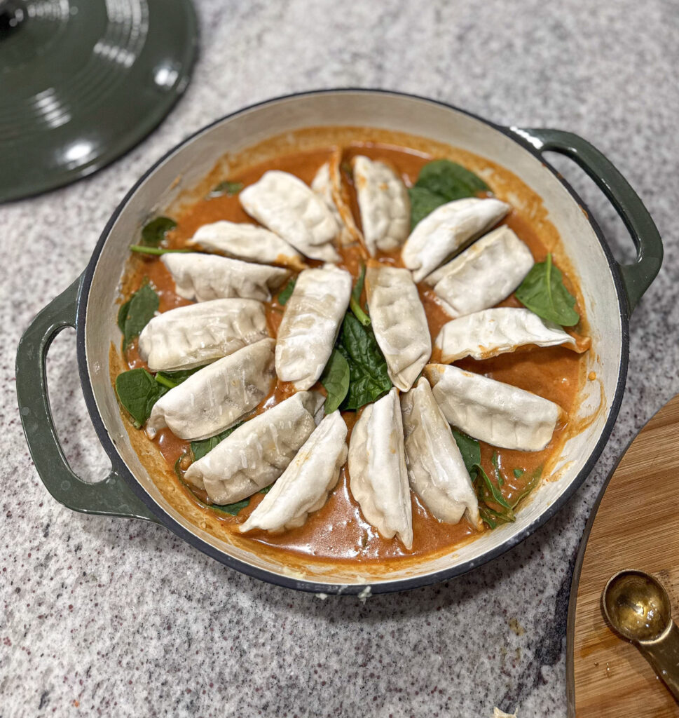 A round green Dutch oven filled with uncooked dumplings arranged in a circle over a creamy red curry sauce with fresh spinach leaves.