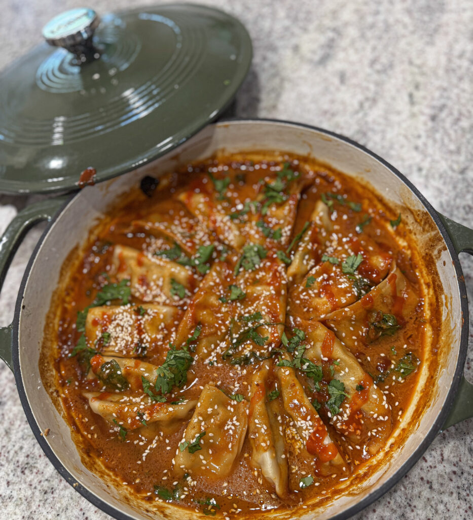 Close-up of golden dumplings baked in a thick red curry sauce, topped with sesame seeds, cilantro, and chili oil in a green Dutch oven.