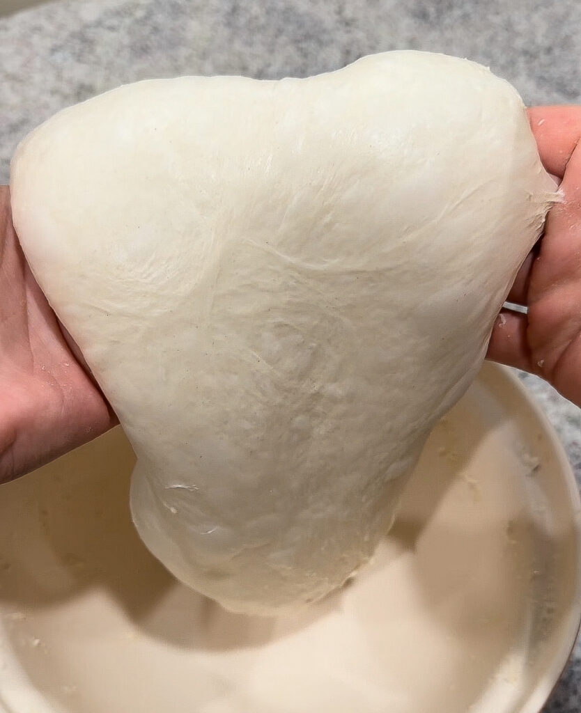 Hands holding up soft sourdough focaccia dough after a coil fold, showing smooth, elastic texture and gluten development.