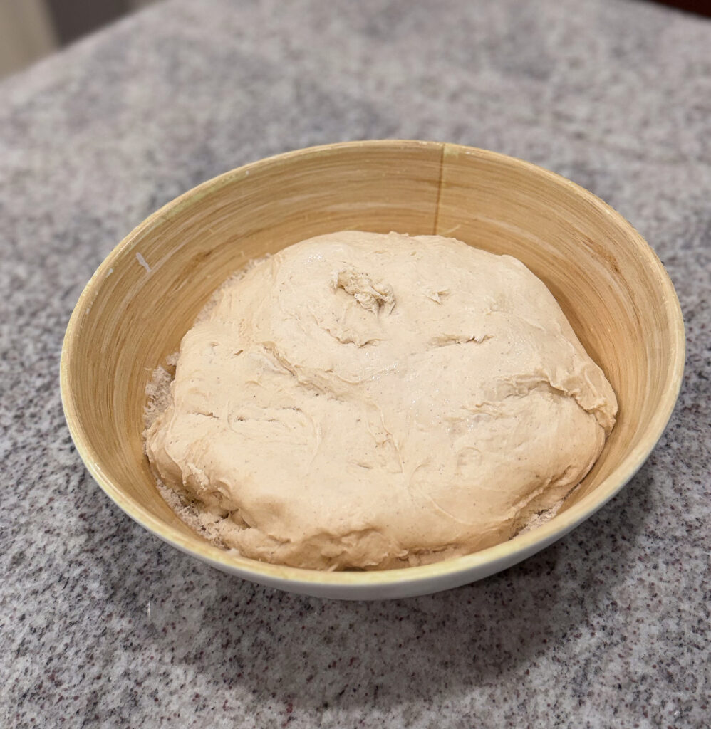 A smooth ball of sourdough donut dough resting in a wooden bowl on a granite countertop, ready for shaping.
