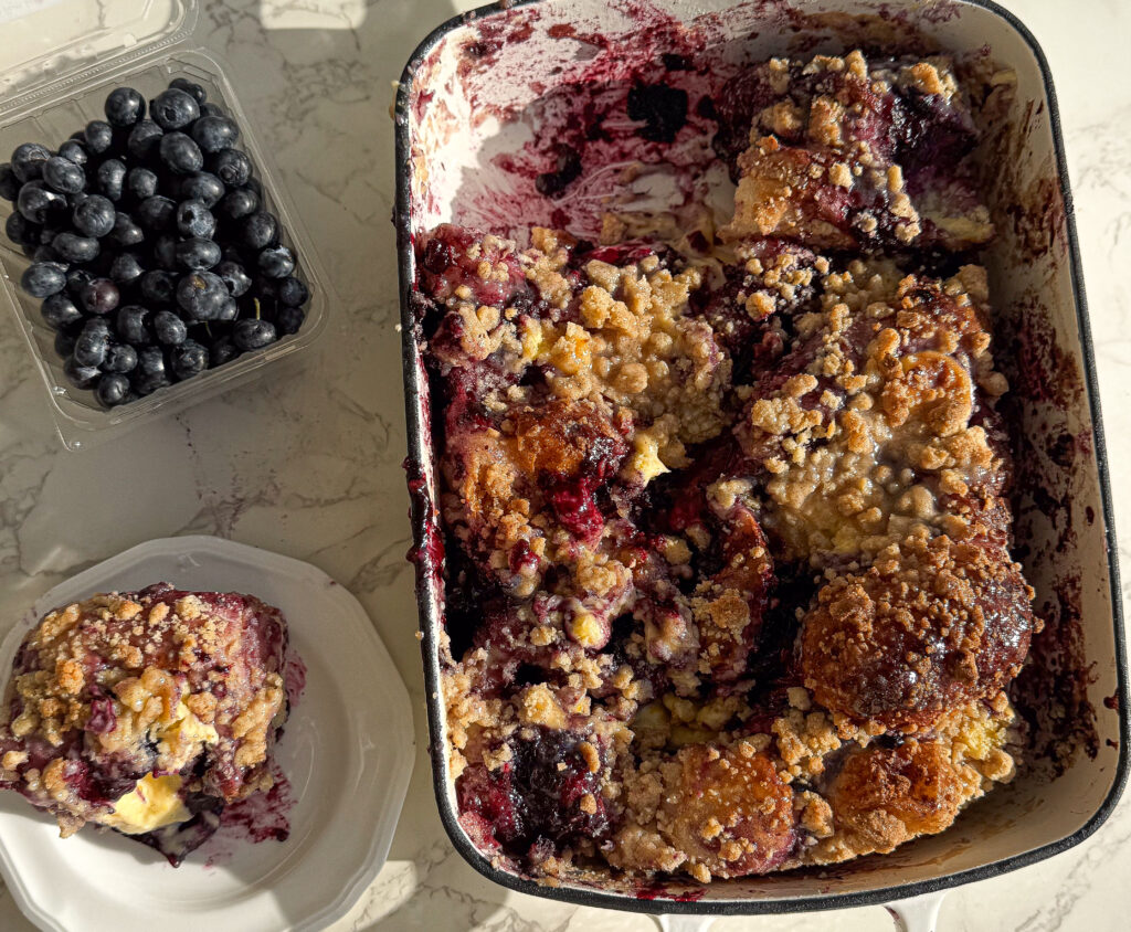 Homemade blueberry sourdough focaccia Danish in a baking pan, topped with crumb streusel and powdered sugar glaze, with a serving on a plate and fresh blueberries on the side.