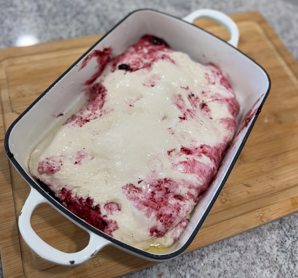 Unbaked sourdough focaccia Danish in a white baking pan, with blueberry compote folded inside the dough and swirls of berry filling showing on top.