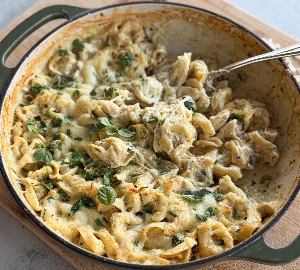 Finished baked chicken Alfredo tortellini topped with melted cheese and fresh herbs in a Dutch oven, sitting on a wooden board with grated parmesan on the side.