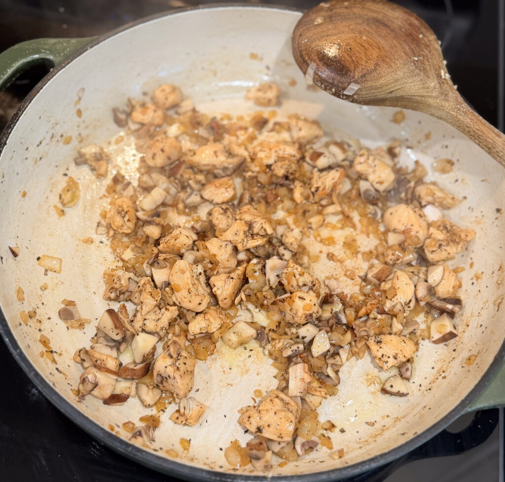 Chopped chicken breast and diced mushrooms sautéing with onions and seasonings in a large enameled cast iron skillet, being stirred with a wooden spoon.
