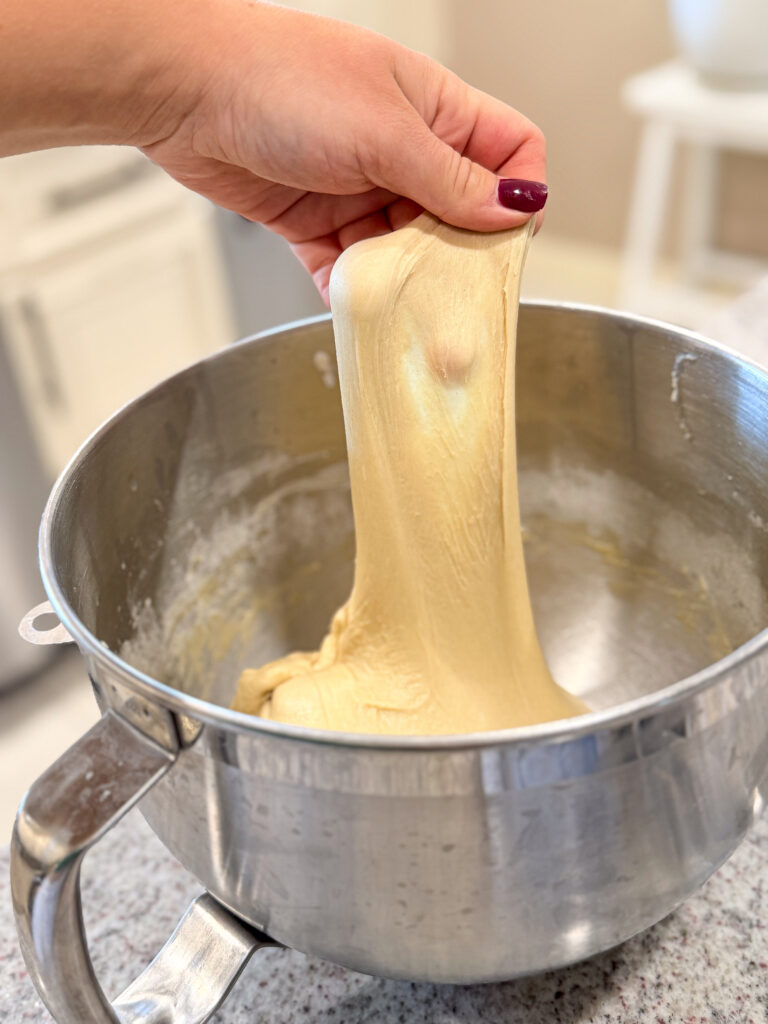 A hand pulling up enriched babka dough from a stand mixer bowl, showing a smooth, stretchy, windowpane-ready texture