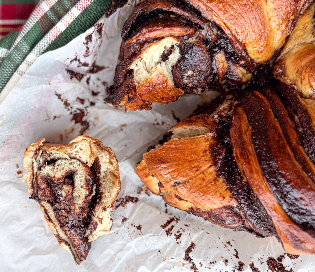 A close-up of a freshly baked sourdough chocolate babka with glossy, golden swirls and a soft chocolate-filled slice pulled away from the loaf on parchment paper.