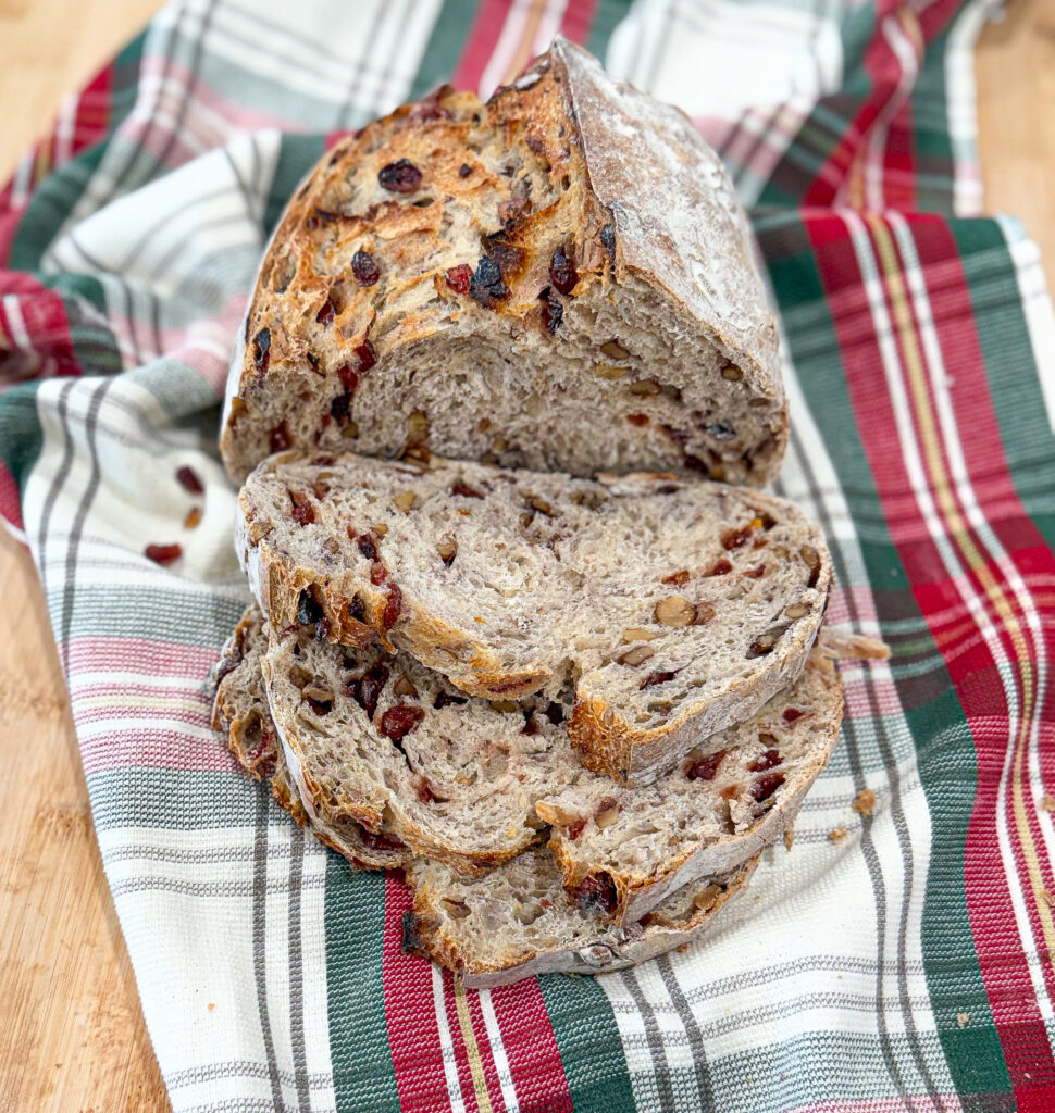 Sliced walnut cranberry sourdough loaf displayed on a festive plaid cloth, showing the golden crust and swirled crumb filled with dried cranberries and chopped walnuts.