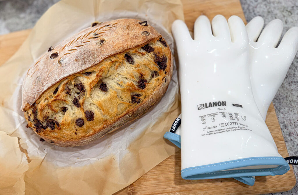 Chocolate chip sourdough croissant bread loaf resting on parchment paper beside a pair of white LANON heat-resistant gloves on a wooden board.