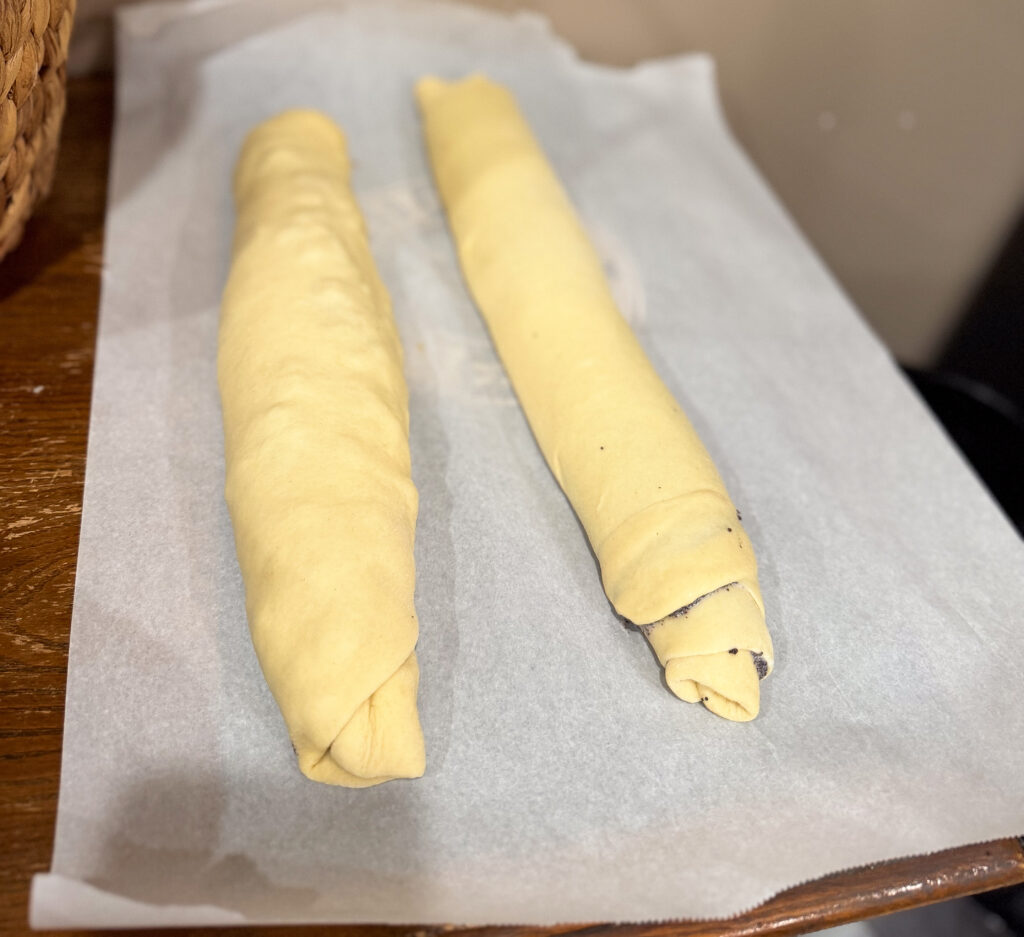 Two shaped vertuta rolls filled with poppy seed mixture resting on parchment paper before final proofing.