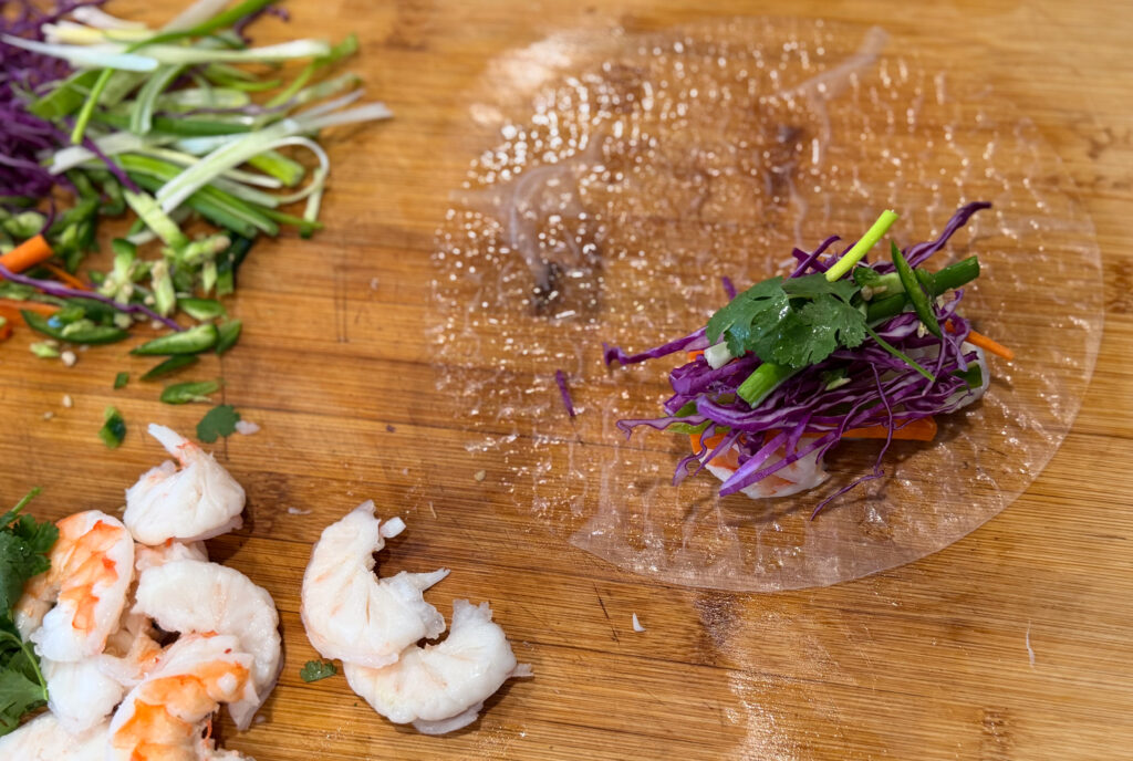 Shrimp spring roll being assembled on rice paper with shredded purple cabbage, carrots, green onions, cilantro, and cooked shrimp on a wooden cutting board.