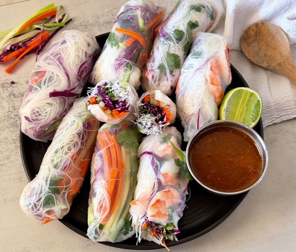 Fresh shrimp spring rolls filled with rice noodles, vegetables, and herbs served on a black plate with peanut dipping sauce and lime wedges.