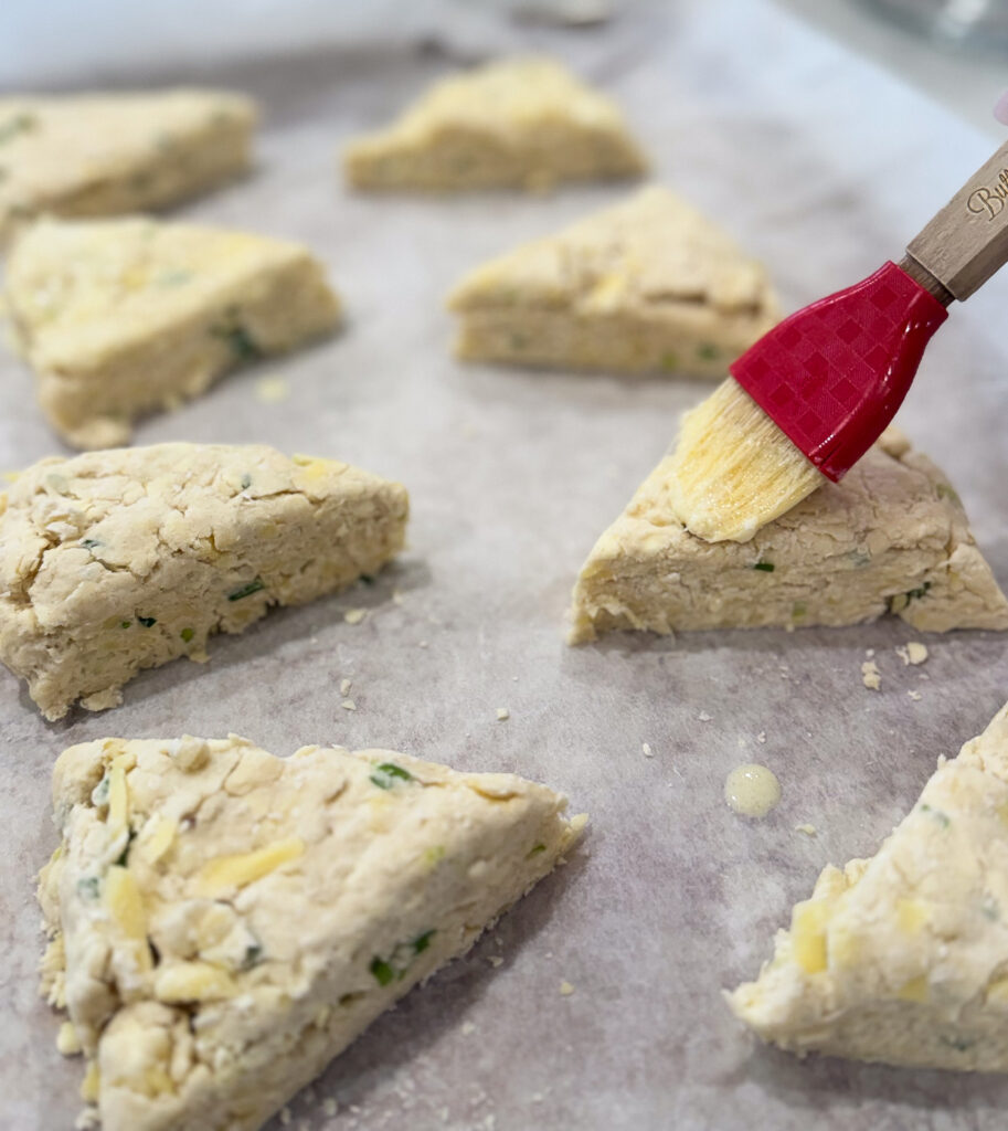IMG_0463 Unbaked triangular sourdough cheese scones on parchment paper as garlic heavy cream wash is brushed over the tops before baking.
