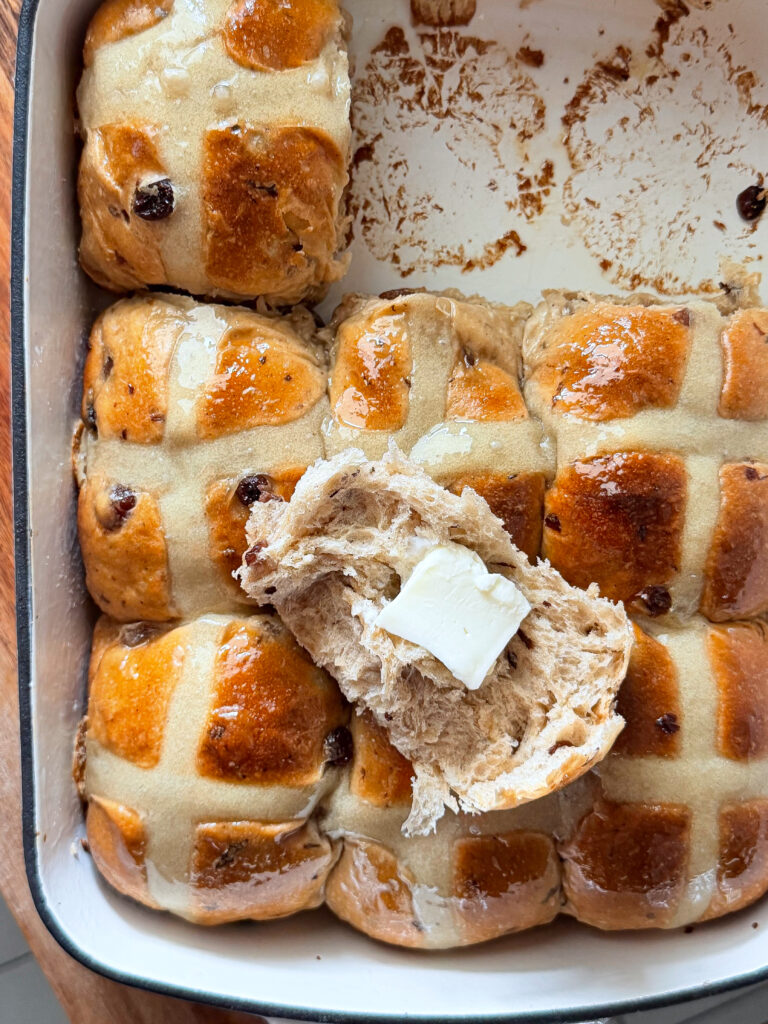 Freshly baked sourdough hot cross buns with golden brown tops, piped crosses, and glossy sugar glaze, one bun torn open with butter inside