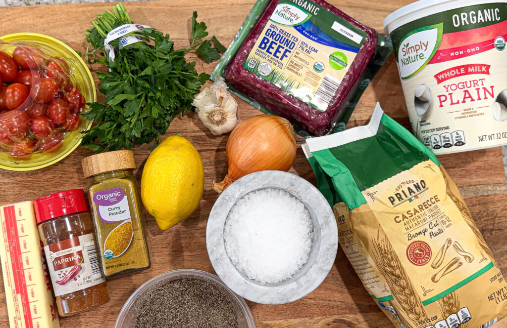 Ingredients for Turkish pasta including ground beef, yogurt, cherry tomatoes, garlic, onion, parsley, lemon, pasta, and spices on a wooden board