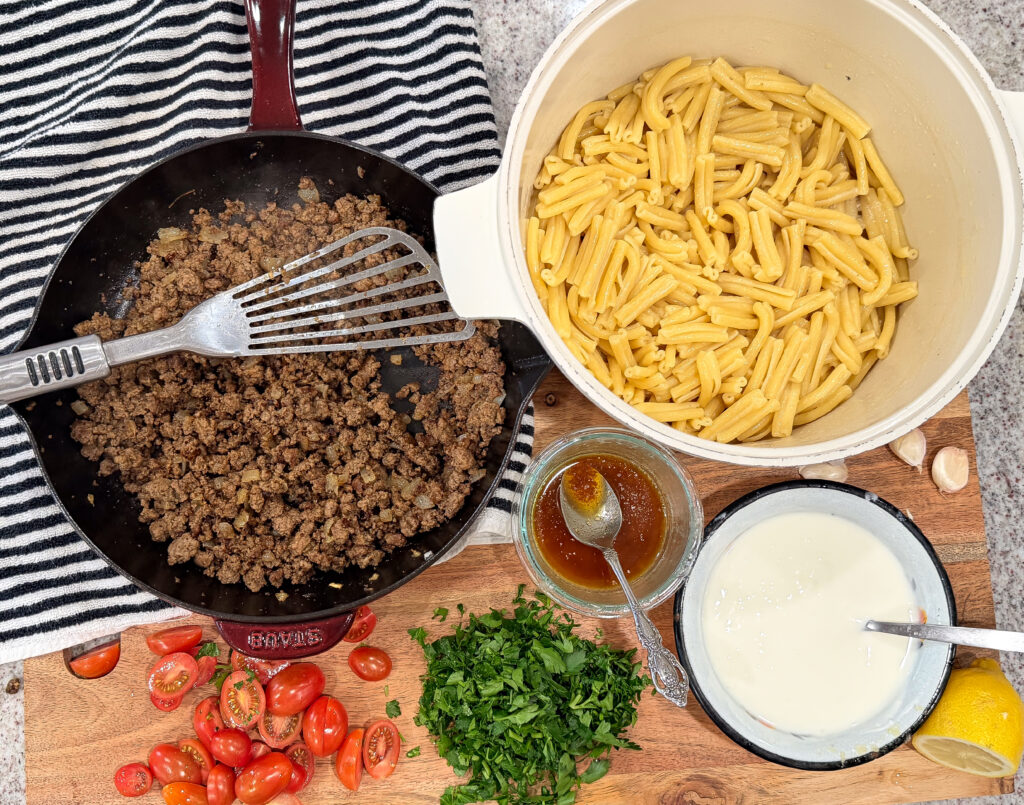 Cooked pasta and seasoned ground beef in a skillet with garlic yogurt sauce, chopped parsley, and tomatoes prepared for Turkish pasta