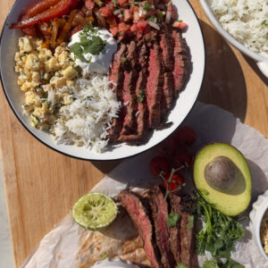 A plate with sliced steak, white rice, sautéed peppers, corn, pico de gallo, and sour cream; nearby are more steak, pico de gallo, avocado, lime, and rice.