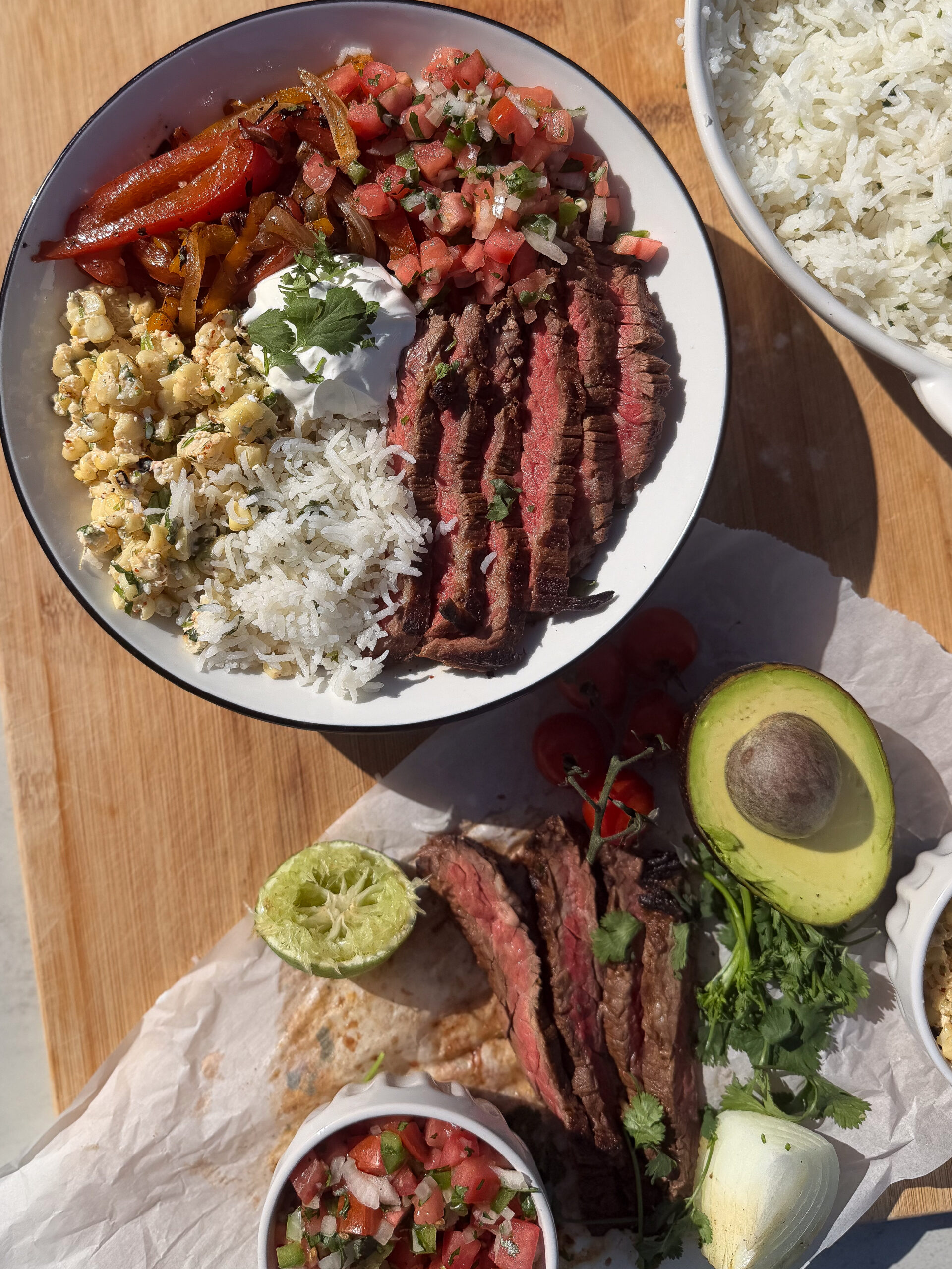 A plate with sliced steak, white rice, sautéed peppers, corn, pico de gallo, and sour cream; nearby are more steak, pico de gallo, avocado, lime, and rice.