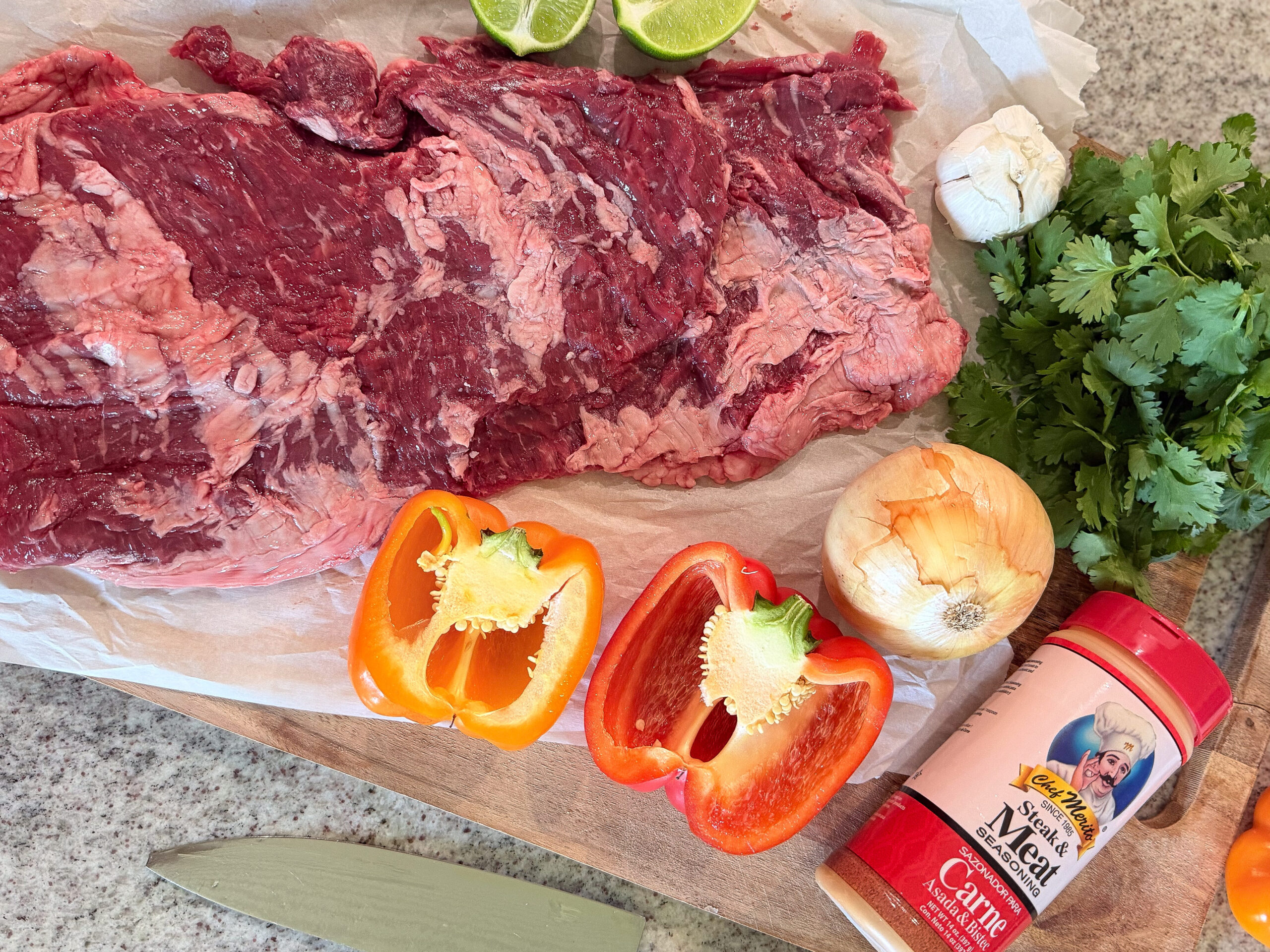 Raw skirt steak on parchment paper with cilantro, garlic, lime halves, onion, red and orange bell peppers, seasoning container, and a knife on a cutting board.