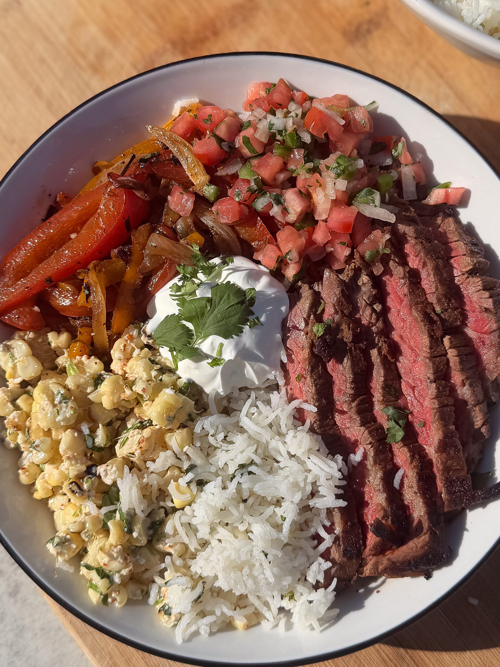 A bowl with sliced grilled steak, white rice, corn salad, sautéed peppers, pico de gallo, and a dollop of sour cream topped with cilantro.