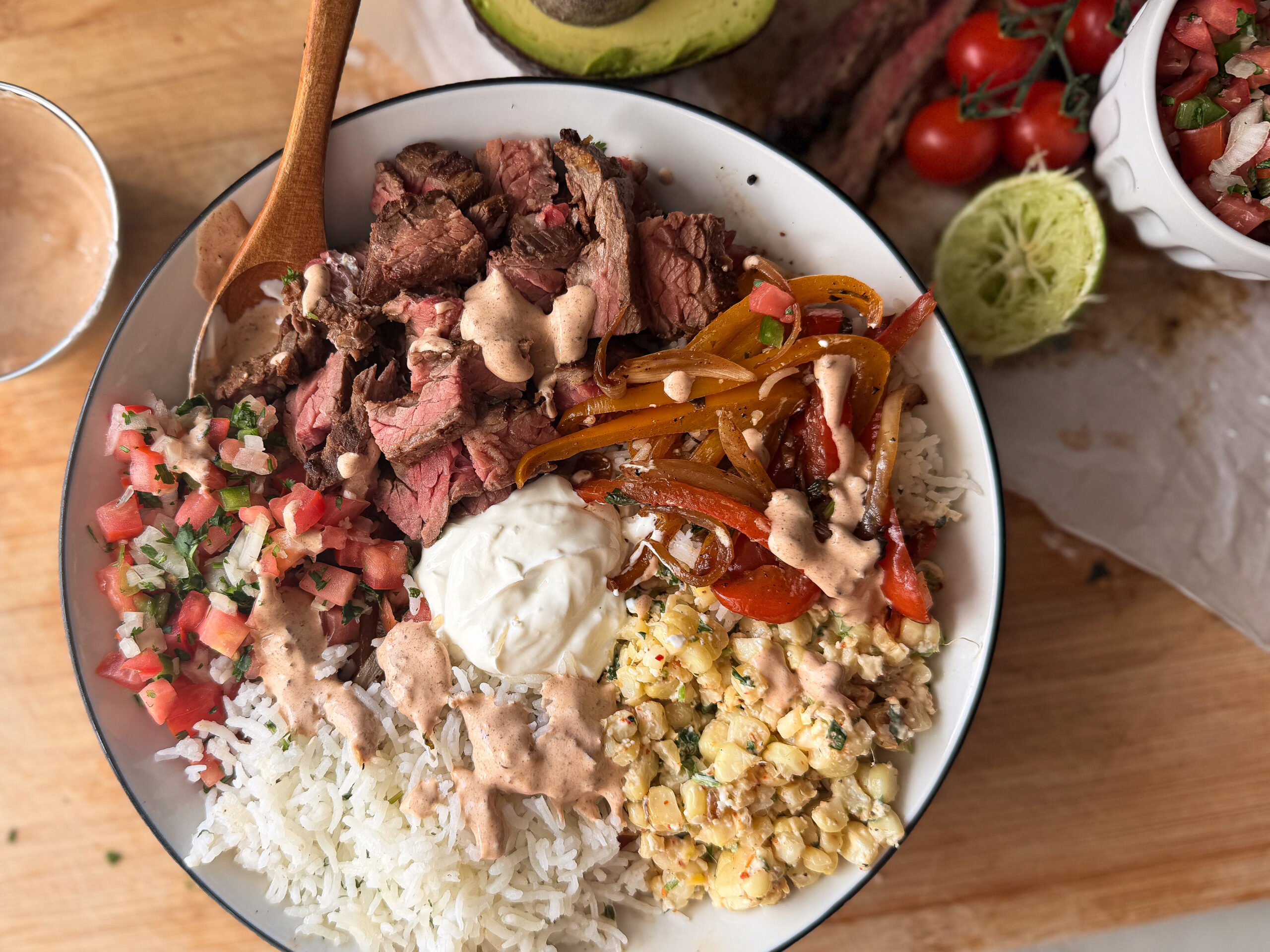 A bowl containing rice, grilled steak slices, sautéed bell peppers, corn, pico de gallo, sour cream, and sauce, with fresh ingredients like avocado and tomatoes nearby.