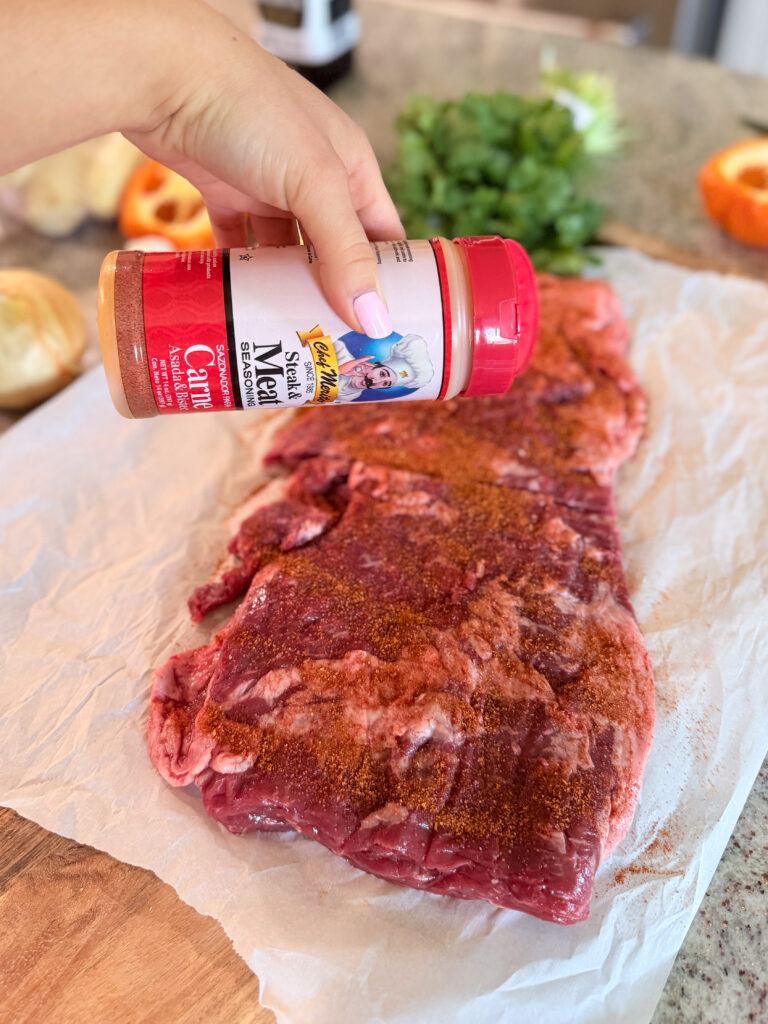 A hand sprinkles meat seasoning onto a raw piece of beef laying on parchment paper, with fresh herbs and vegetables in the background.