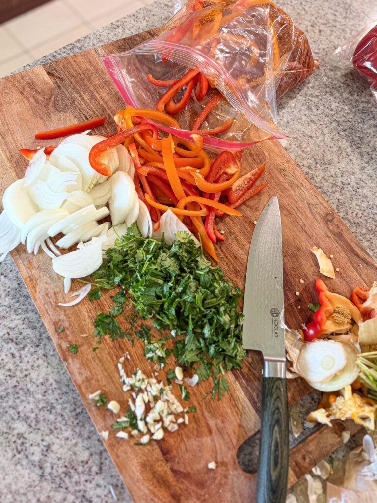 A wooden cutting board with sliced onions, chopped cilantro, minced garlic, and sliced red and orange bell peppers beside a chef’s knife and vegetable scraps.