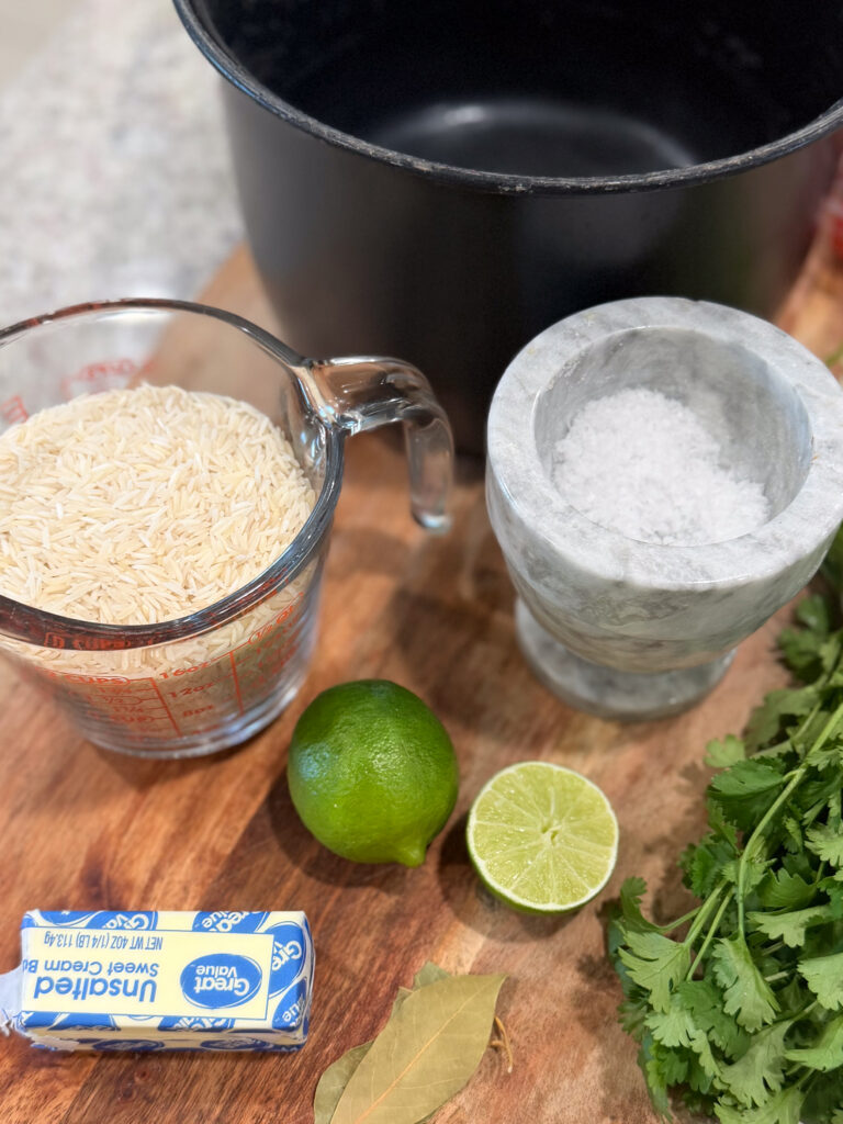 A cutting board with a measuring cup of uncooked rice, butter, a halved lime, bay leaf, cilantro, a mortar with salt, and a black pot in the background.
