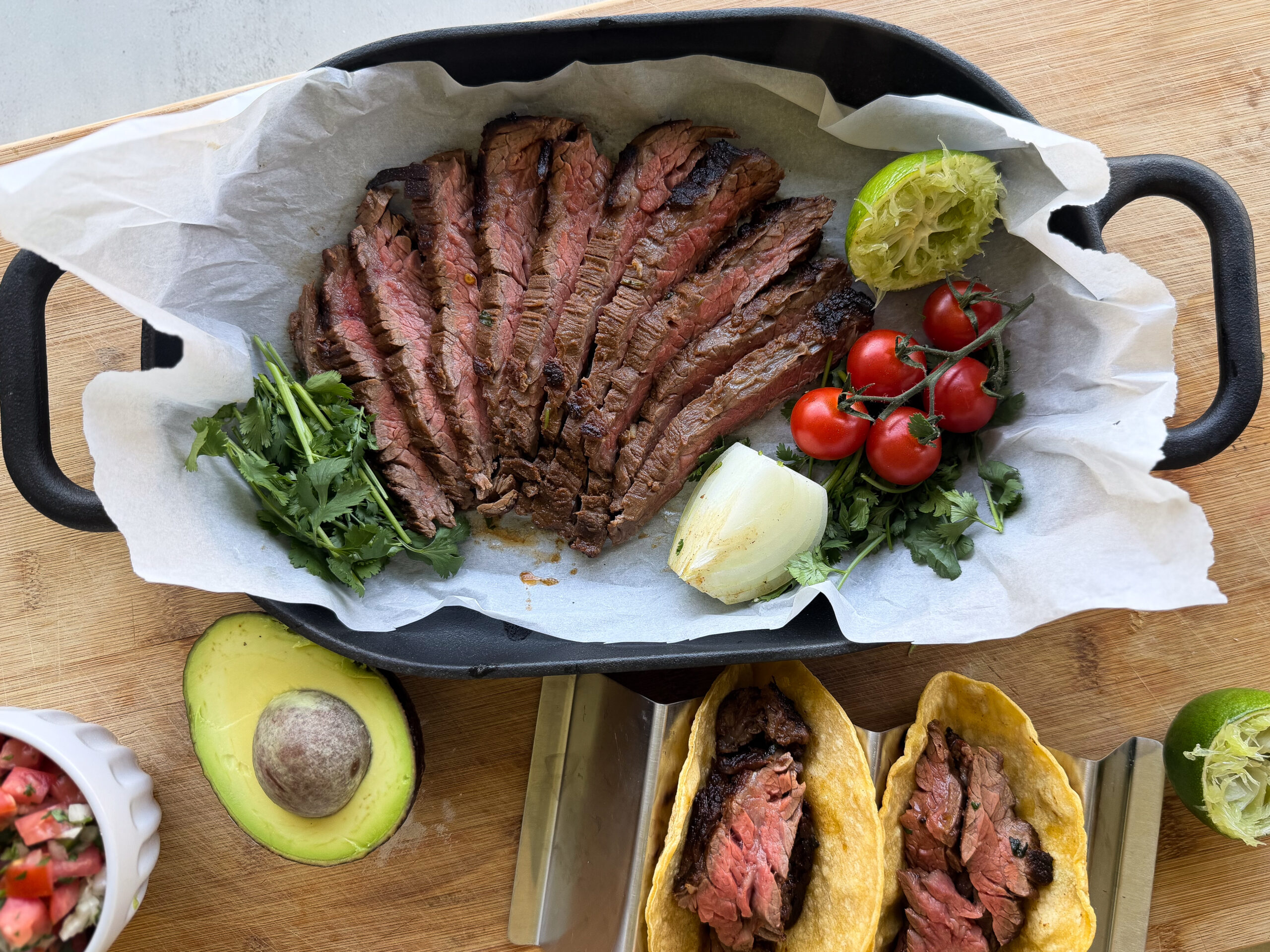 Sliced grilled steak in a black pan with parchment paper, garnished with greens, cherry tomatoes, grilled onion, and lime; tacos with steak in the foreground, avocado and salsa on the side.