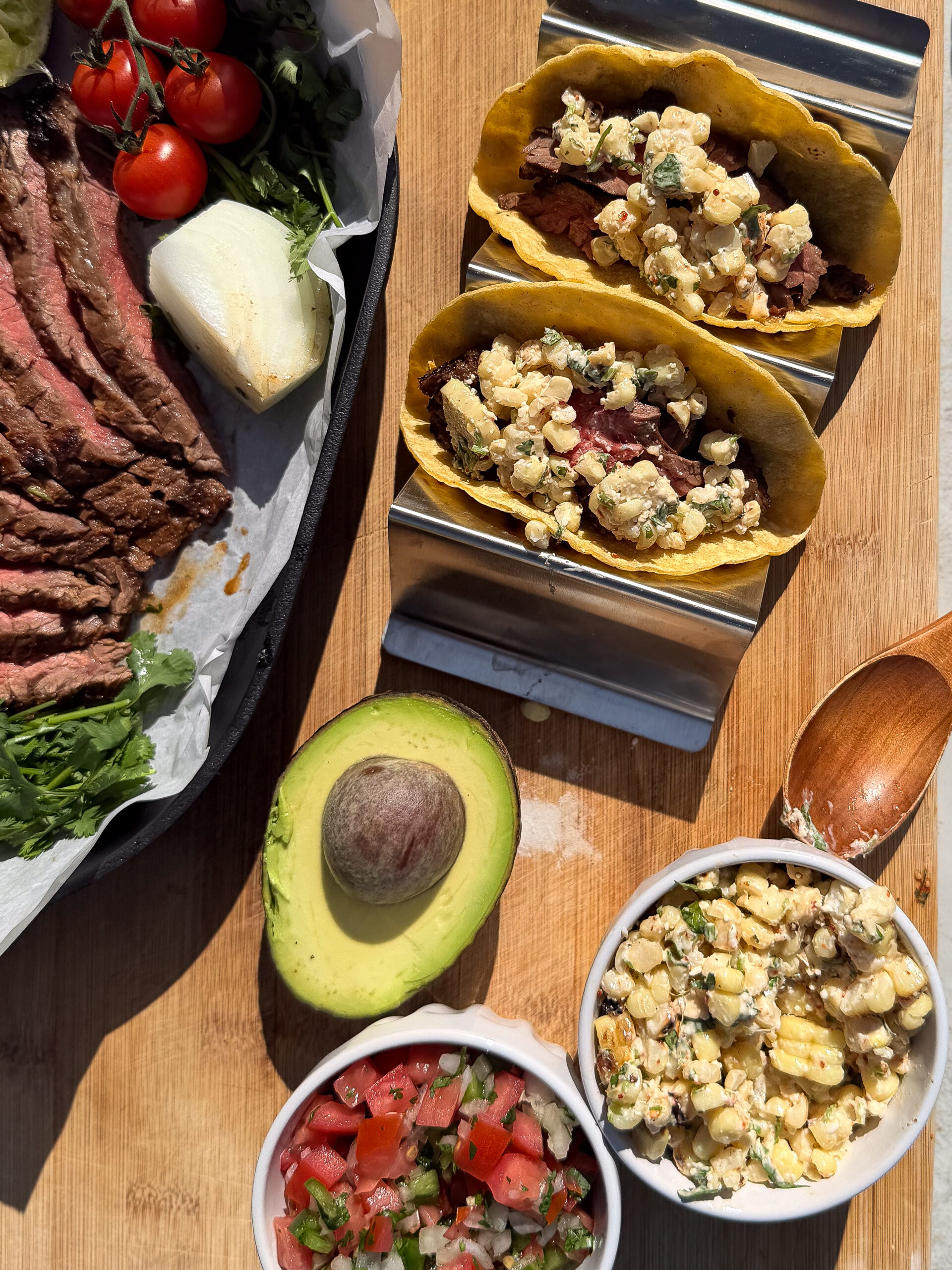 Three steak and corn tacos are displayed on a wooden surface, accompanied by sliced steak, cherry tomatoes, half an avocado, pico de gallo, and a bowl of corn salad.