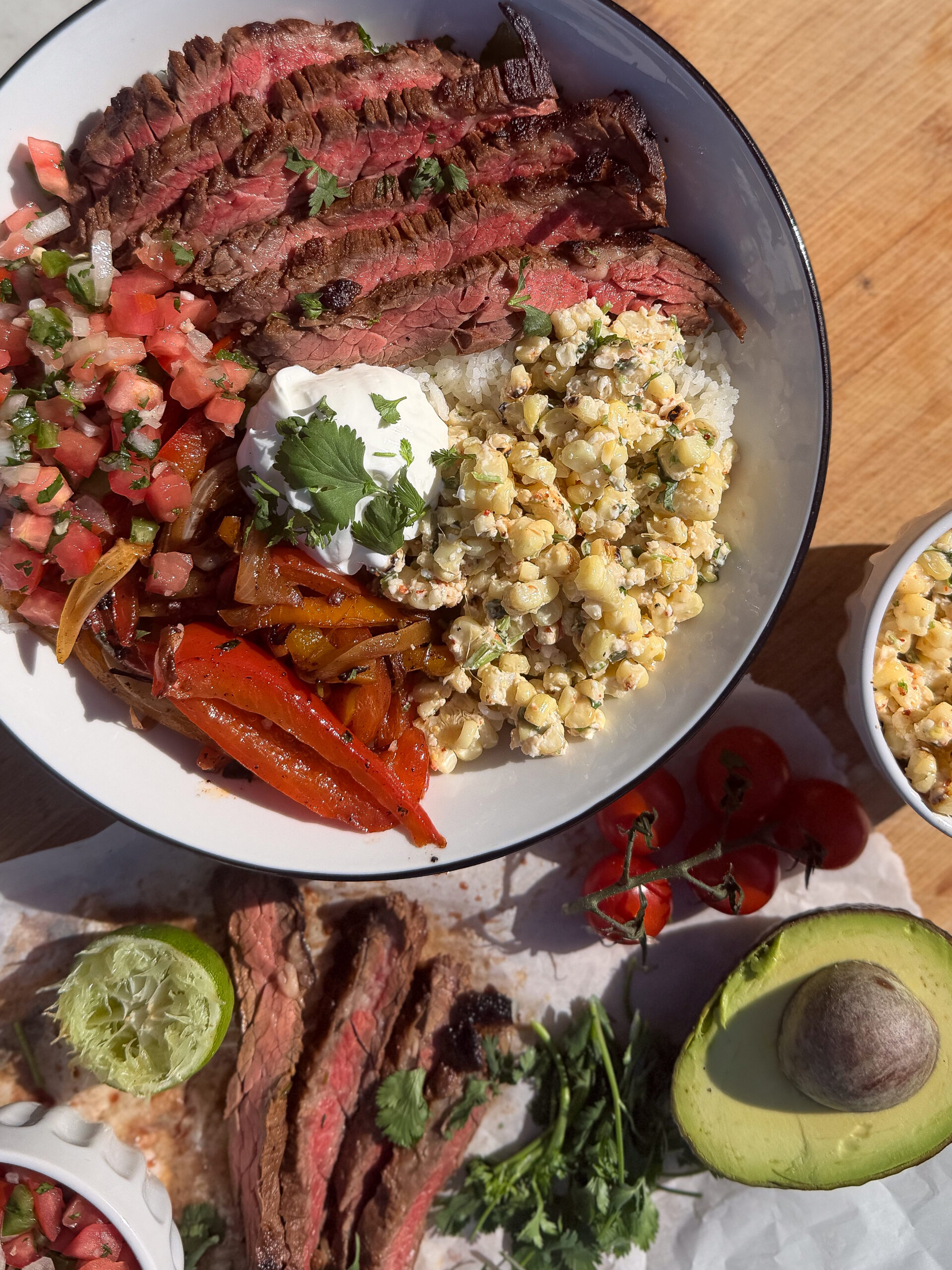 A bowl with sliced grilled steak, corn, diced tomatoes, sautéed red peppers, rice, and a dollop of sour cream, with avocado, cilantro, lime, and extra steak on the side.