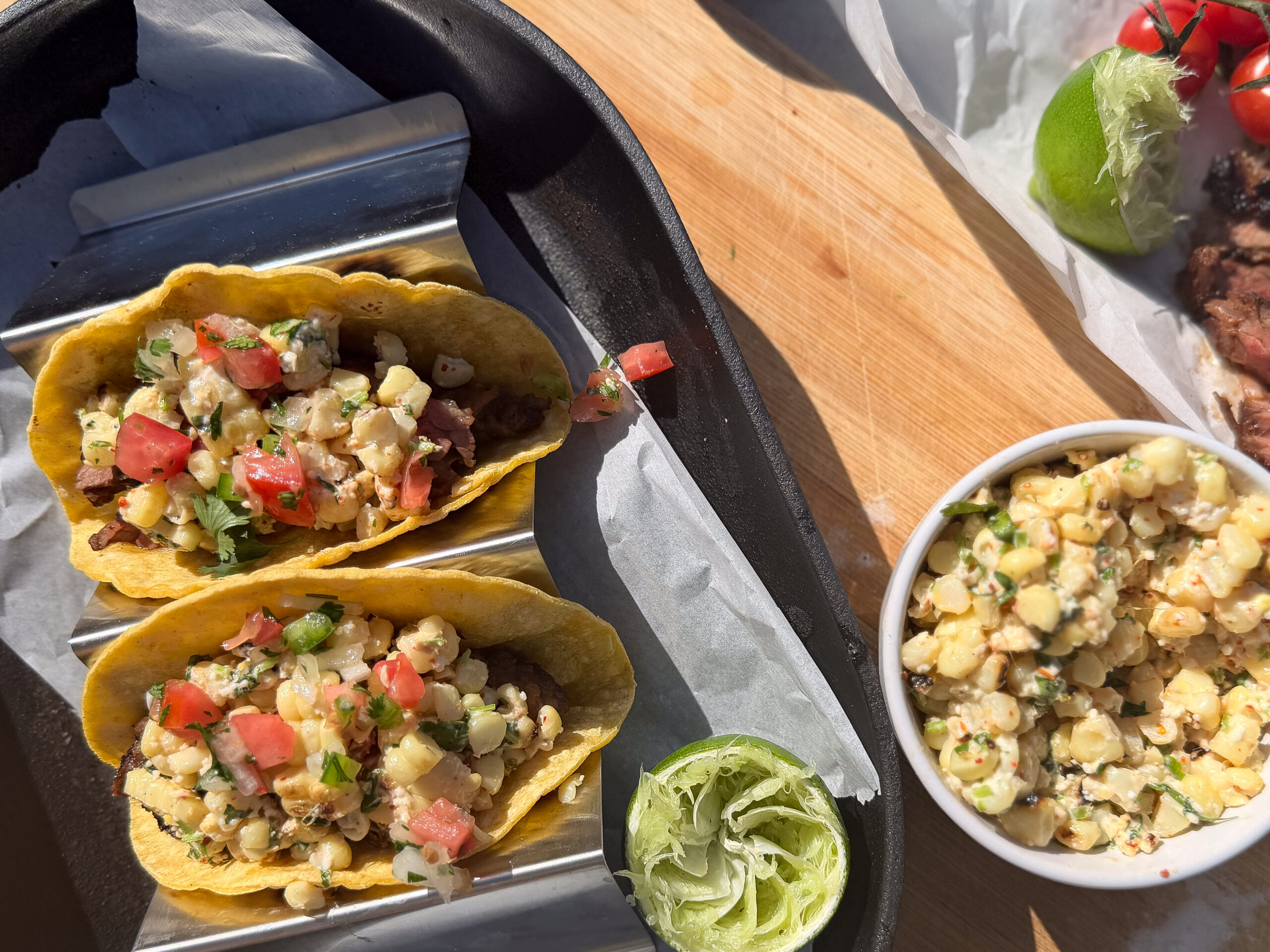 Two tacos with corn salsa, diced tomatoes, and cilantro on a tray with lime slices. A bowl of corn salsa and a lime are on the side.