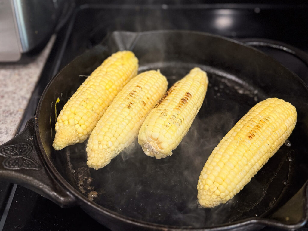 Four ears of corn are being roasted in a black cast iron skillet, with some grill marks and visible steam rising from the pan.