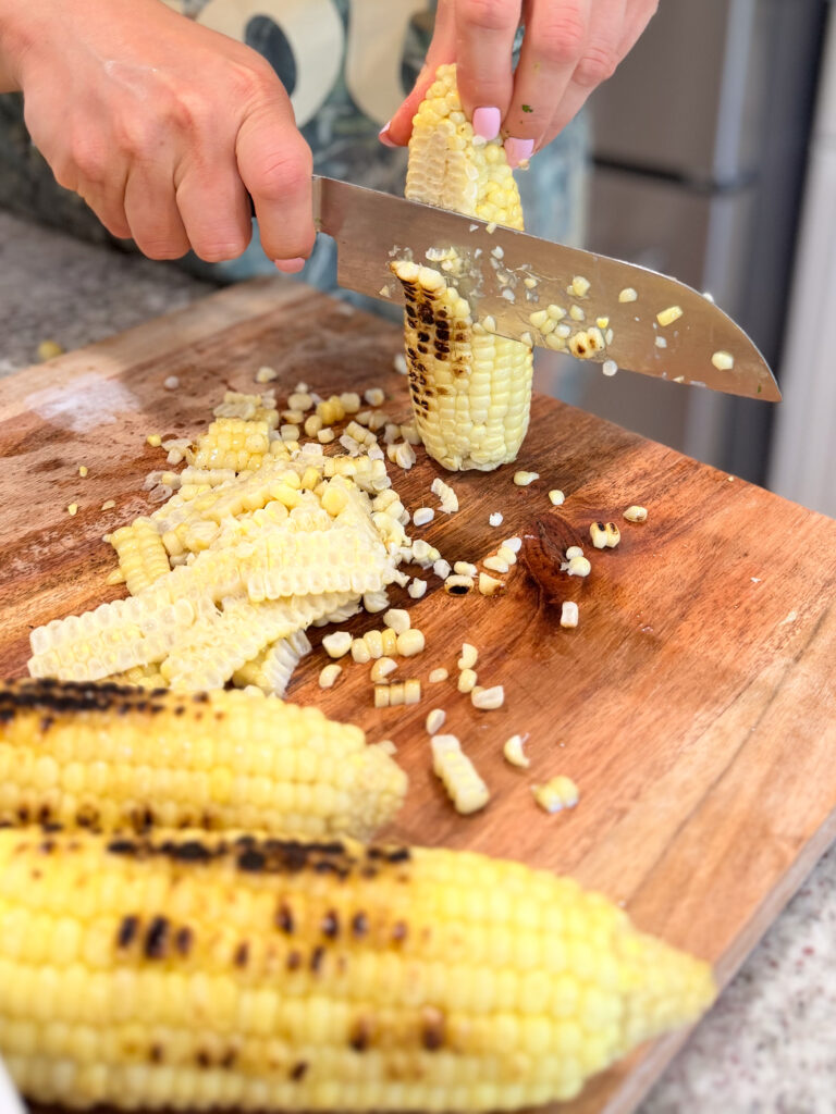 Person cutting grilled corn kernels off the cob with a knife on a wooden cutting board, with extra corn cobs nearby.