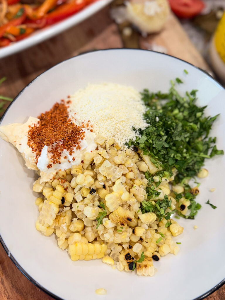 A bowl containing grilled corn kernels, chopped cilantro, grated cheese, mayonnaise, and chili powder, prepared for mixing.