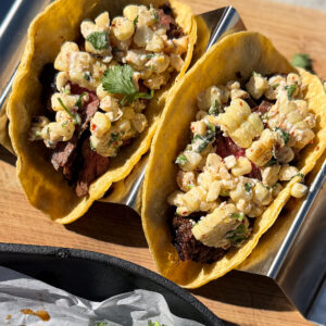 Two tacos filled with grilled meat and a corn salad garnish, served in a metal taco holder on a wooden surface.