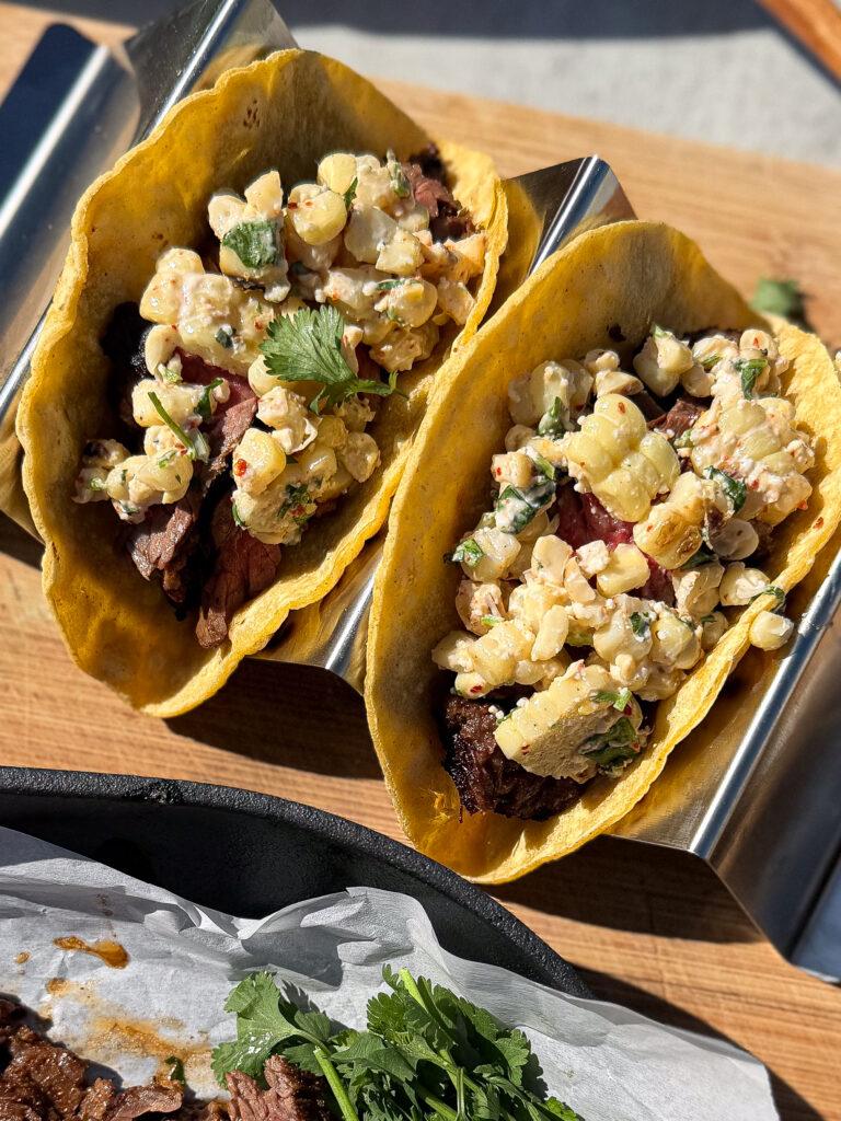 Two tacos filled with grilled meat and a corn salad garnish, served in a metal taco holder on a wooden surface.