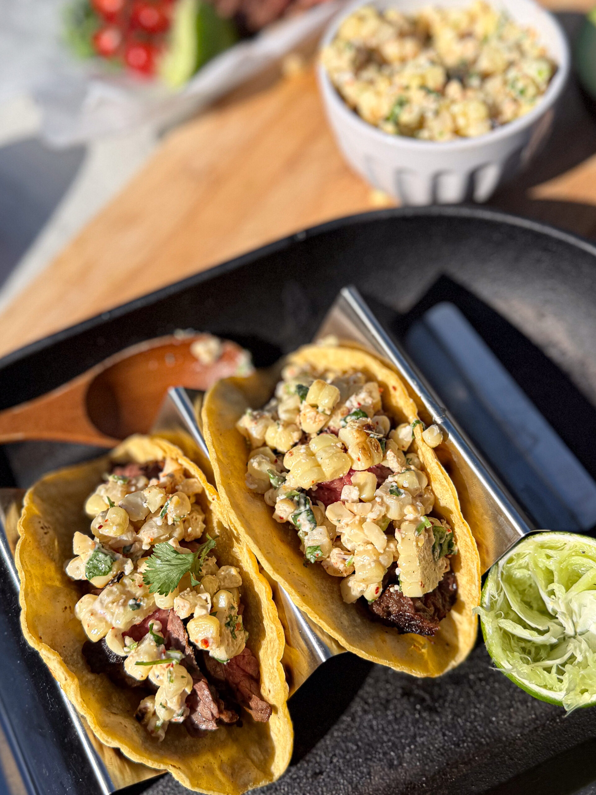 Two tacos filled with grilled meat and corn salad are served on a metal taco holder with a halved lime, and a bowl of corn salad is in the background.
