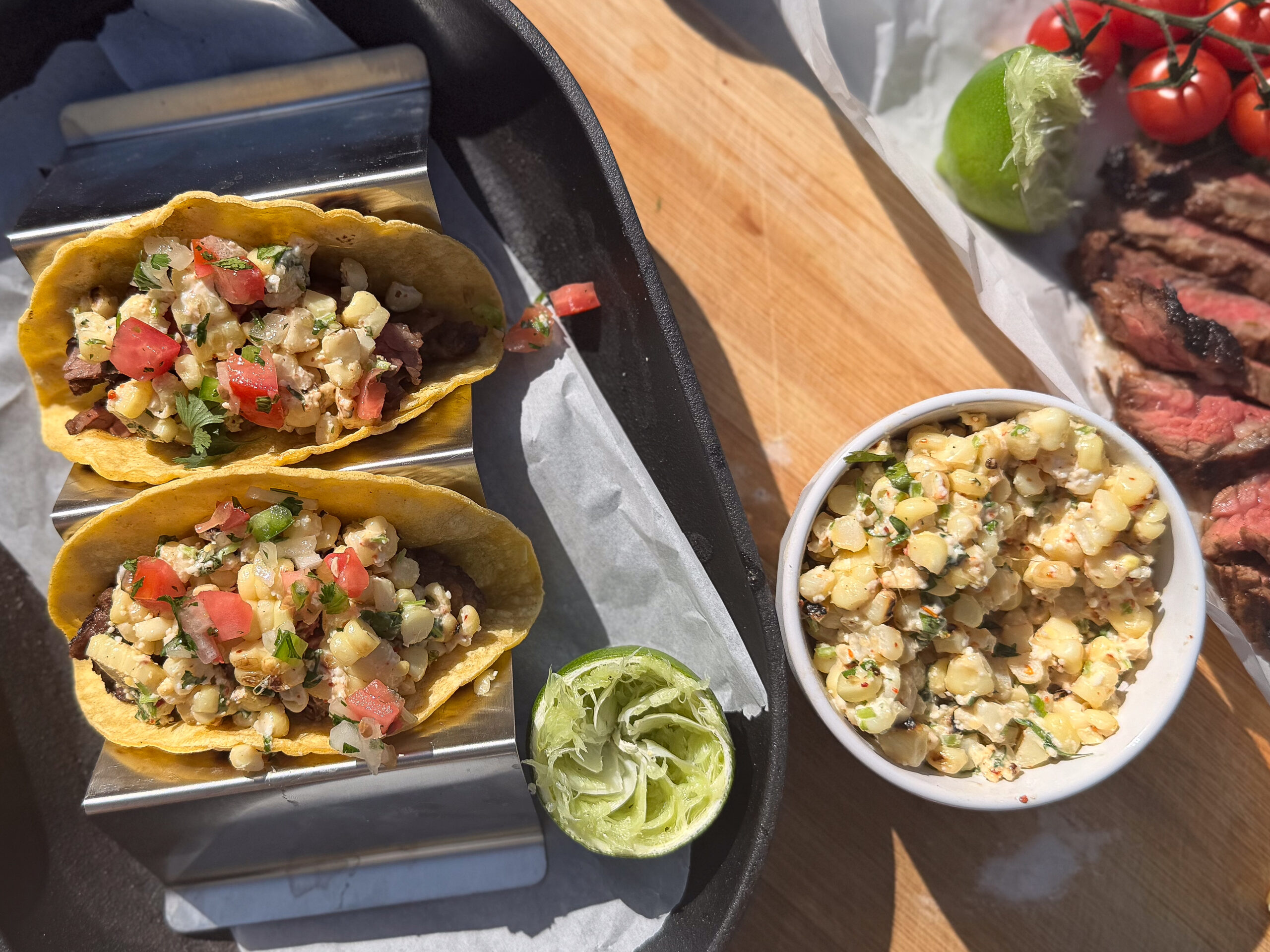 Two tacos filled with corn salsa on a metal holder, a bowl of corn salsa, grilled meat, tomatoes on the vine, and a halved lime on a wooden surface.