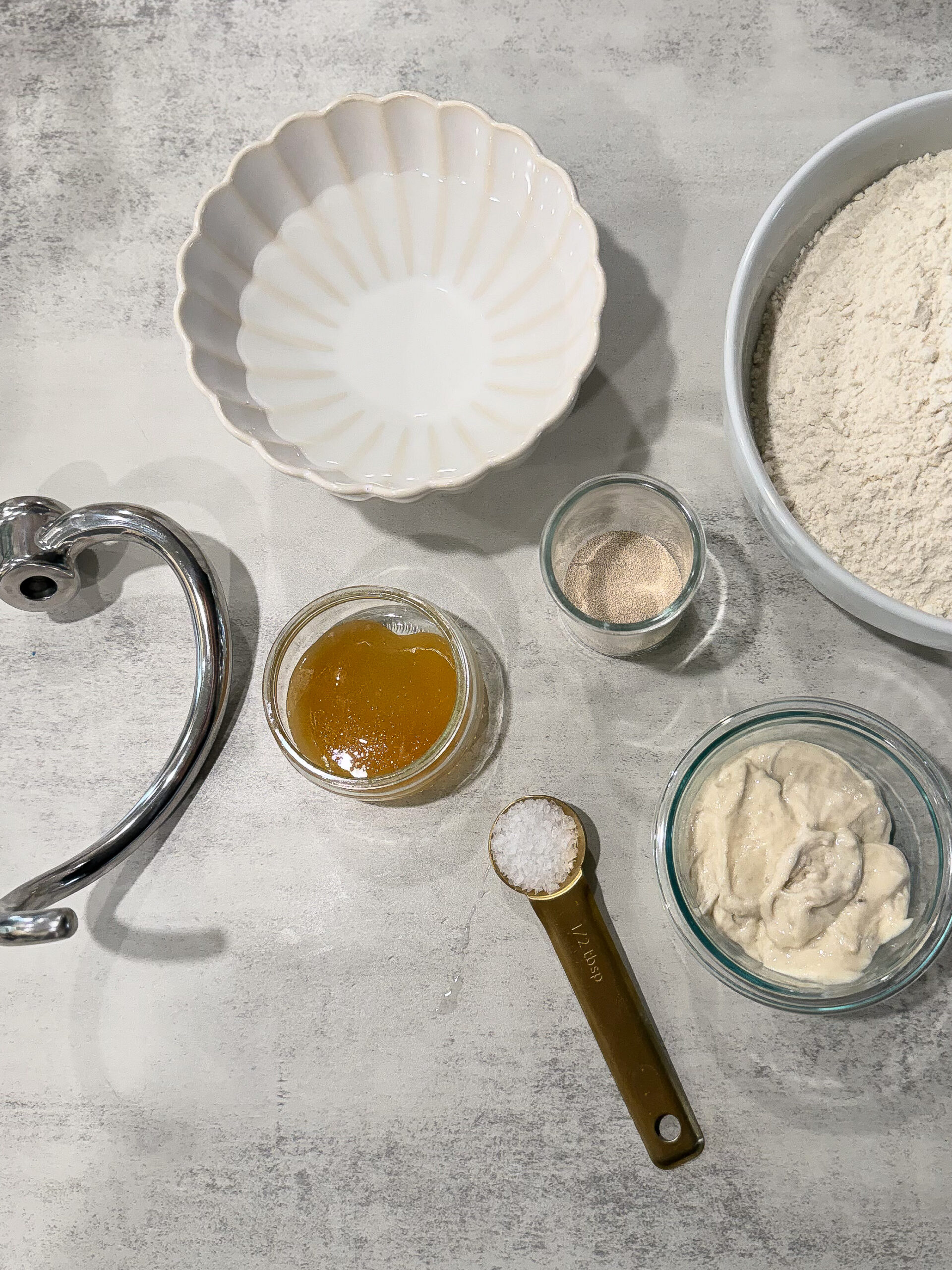 Overhead view of baking ingredients on a countertop: flour, salt, yeast, honey, sourdough starter, an empty bowl, and a mixer dough hook.