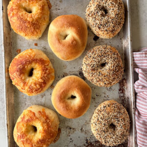 A metal baking tray holds seven assorted bagels: three cheese-topped, two plain, and two everything bagels, next to a folded red and white towel.
