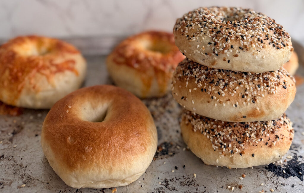 A tray with three everything bagels stacked on each other, two cheese bagels in the background, and one plain bagel in the foreground.