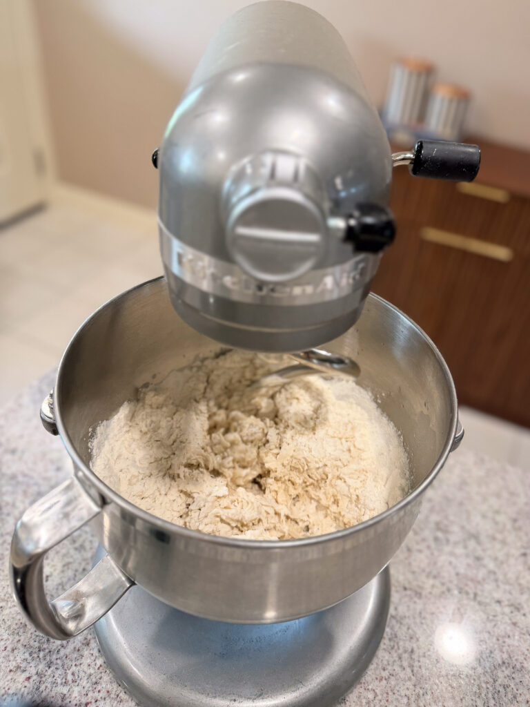 A stand mixer with a dough hook attachment is mixing flour and other ingredients in a metal bowl on a kitchen counter.