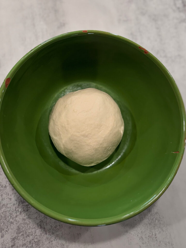 A ball of dough sits in the center of a green bowl on a light-colored surface.